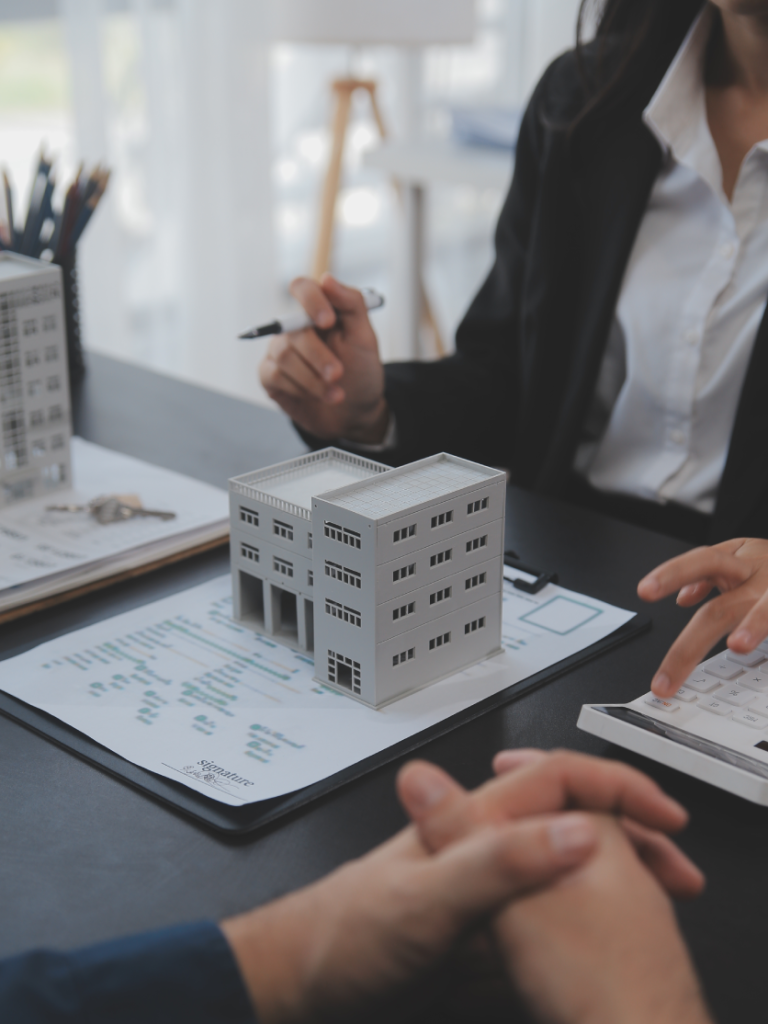 "Una profesional de bienes raíces explicando los detalles de un proyecto utilizando una maqueta de un edificio de apartamentos, una calculadora y documentos de contrato frente a un cliente."