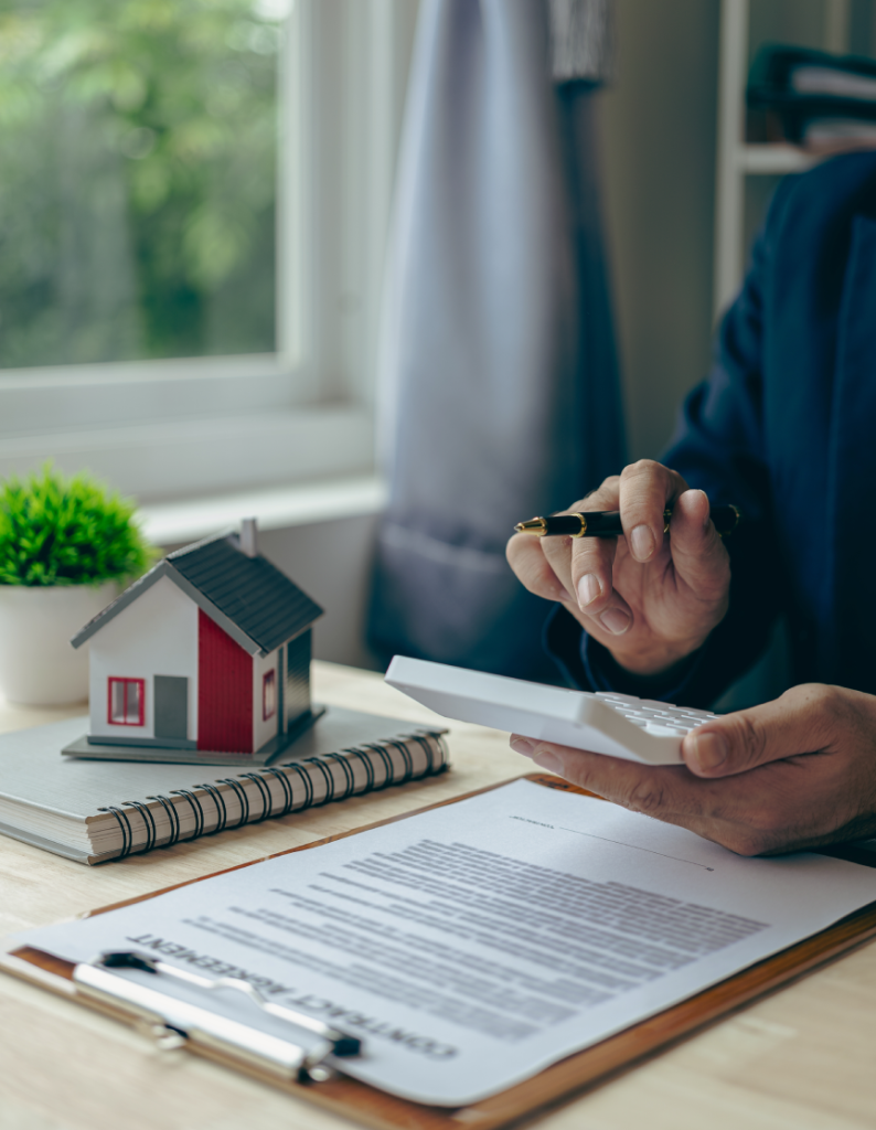 "Primer plano de dos personas dándose la mano sobre una mesa con dos maquetas de casas pequeñas y un contrato, simbolizando un acuerdo inmobiliario exitoso."